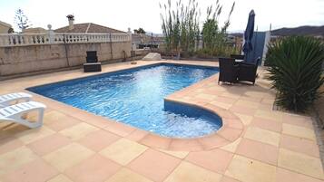 Piscina rectangular con borde curvo, rodeada por un patio empedrado. Hay dos sillones blancos y una pequeña mesa con sillas.