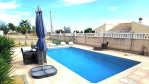 A rectangular swimming pool with a tiled deck, surrounded by lounge chairs and a covered umbrella.