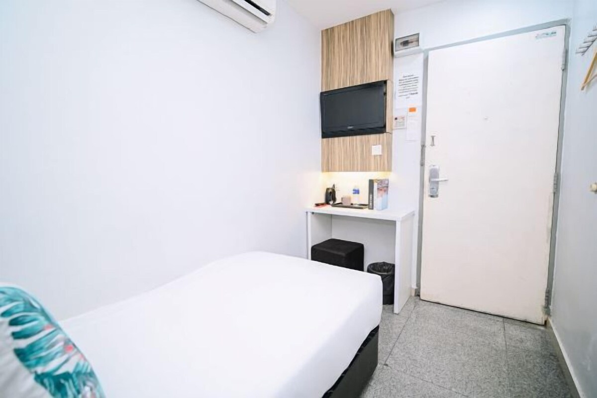 A hotel room with a bed, a TV mounted on a wooden panel, a desk with a chair, and a door.