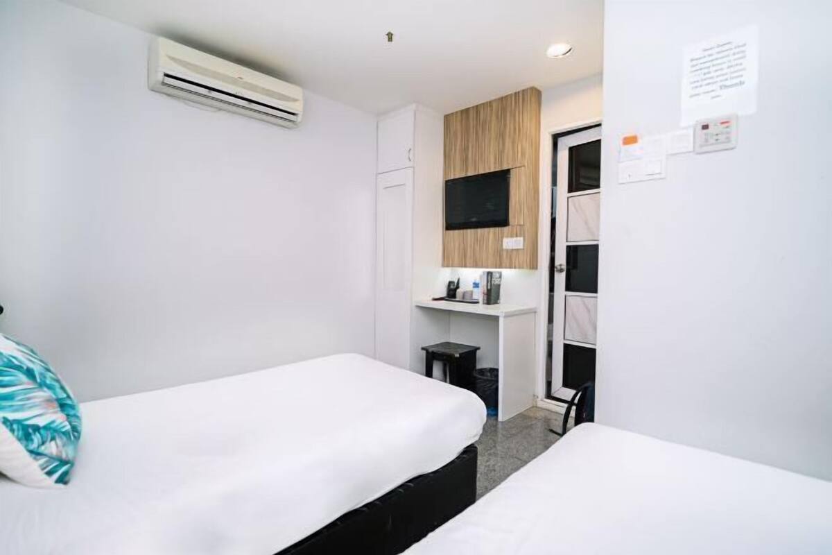 A small hotel room with two single beds, a white air conditioning unit, a wooden panel wall, a small desk with a chair, and a door leading to a bathroom.
