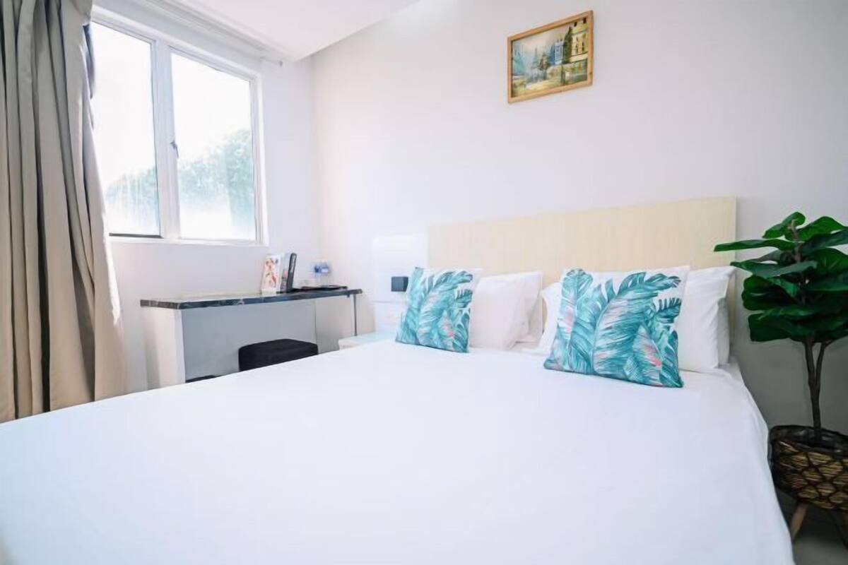 A hotel room with a bed, a desk, a window, and a painting on the wall.
