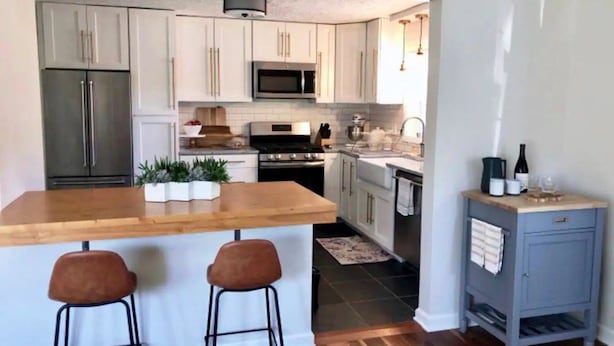 A modern kitchen with white cabinets, stainless steel appliances, a wooden island with bar stools, and a small blue cabinet with wine bottles. - Caitlin's Cottage - Peaceful 2BR Retreat in North Marion (Marion)