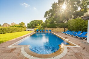 Una piscina rectangular con agua azul, rodeada por un solárium con baldosas. Hay sillas reclinables azules a lo largo del borde de la piscina.
