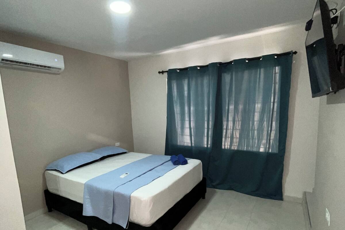 A bedroom with a bed, a ceiling fan, an air conditioning unit, and a window with curtains.