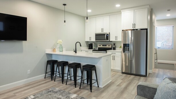 A modern kitchen with white cabinets, a stainless steel refrigerator, a microwave, and a built-in oven. There are four black bar stools at the island. - Pleasant Stay Place - New Build/Zion/Sand Hollow (Hurricane)