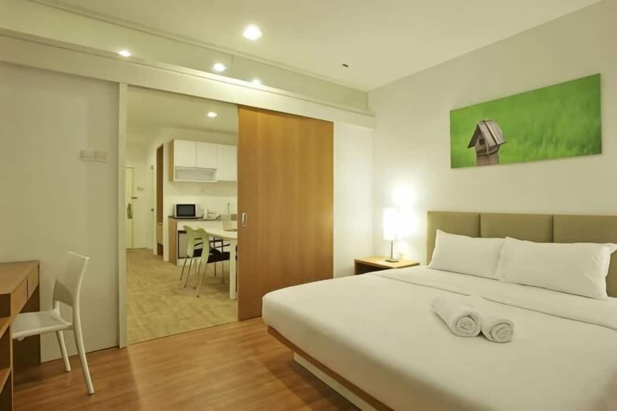 A hotel room with a bed, a desk, a chair, a nightstand, a painting, and a view into the kitchen area.