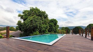 A rectangular swimming pool with a wooden deck surrounding it, set against a backdrop of lush greenery and distant mountains. - The Brompton's Luxury Apt (Kingston)