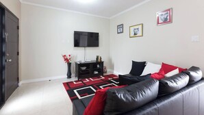 A living room with a black leather sofa, a flat-screen TV mounted on the wall, a black entertainment unit, and a red and black patterned rug. - The Brompton's Luxury Apt (Kingston)