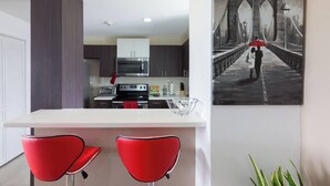 A modern kitchen with dark cabinets, a white countertop, and red chairs. A painting of a bridge is on the wall. - The Brompton's Luxury Apt (Kingston)