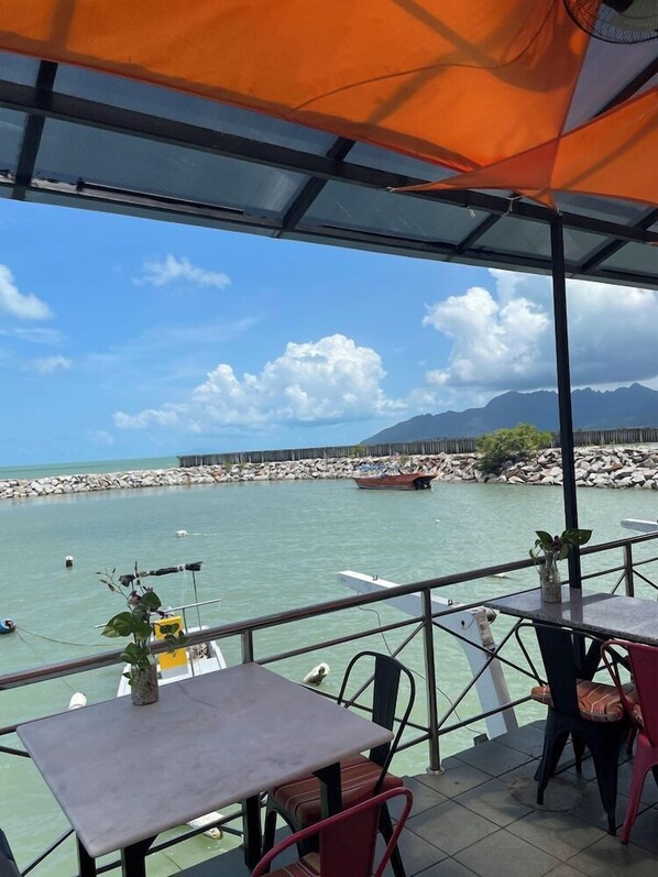 A terrace with tables and chairs overlooking a calm sea, a rocky breakwater, and a mountain in the distance.