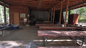 An outdoor covered picnic area with wooden benches and picnic tables, a wooden door, and a kitchen area in the background.