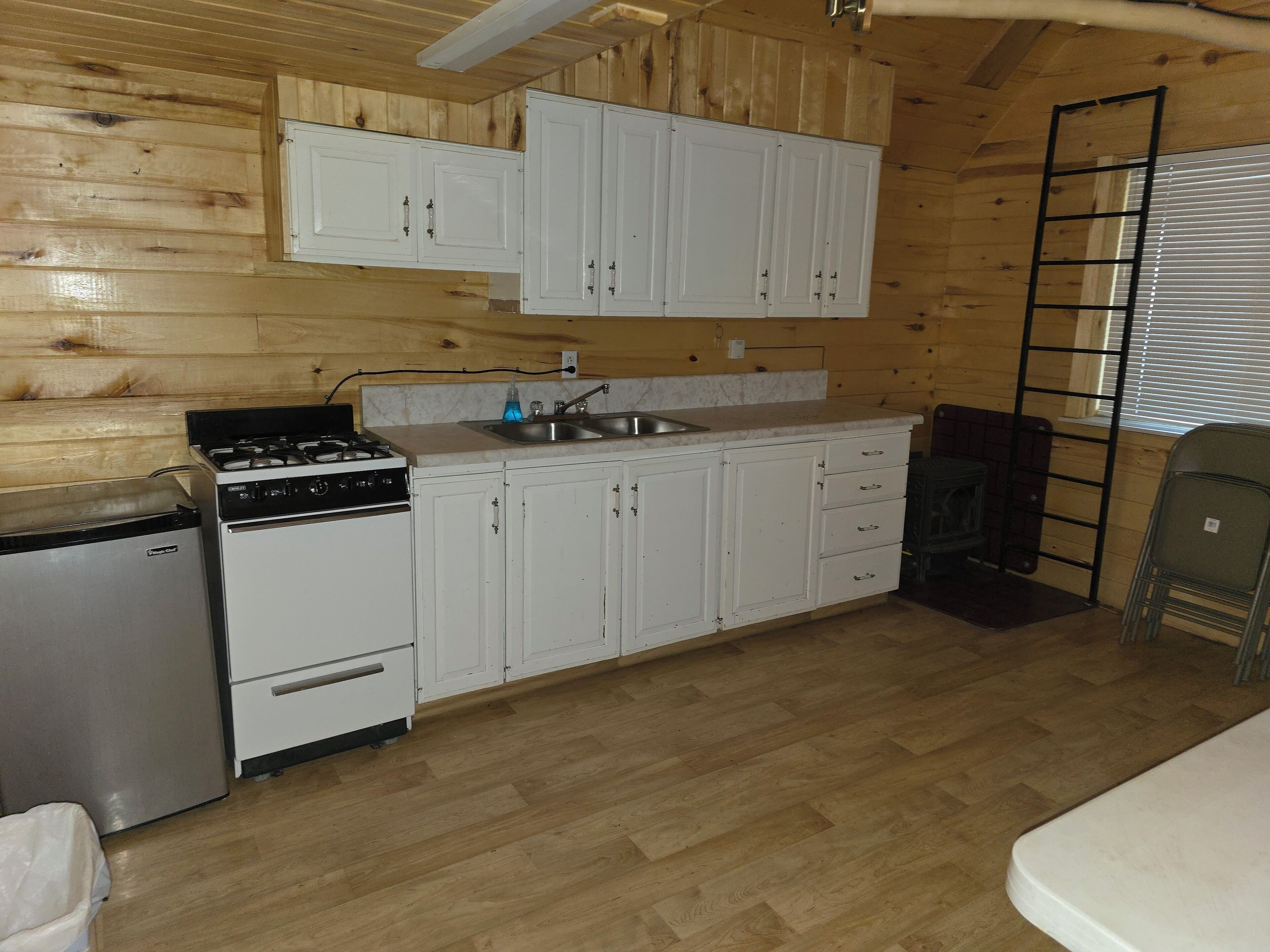 A compact kitchen with wooden walls, white cabinets, a stove, a sink, and a small refrigerator.