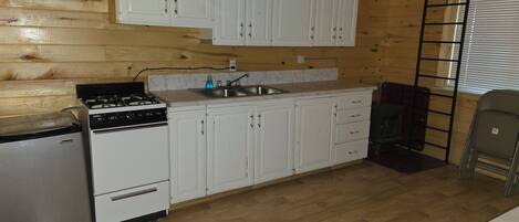 A compact kitchen with wooden walls, white cabinets, a stove, a sink, and a small refrigerator.