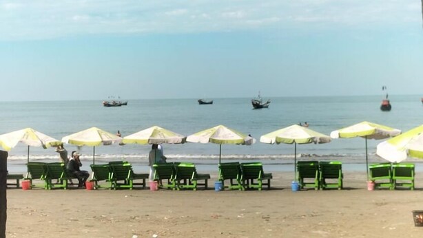 Een strand met rijen groene stoelen en gele parasols, boten in het water, en een heldere lucht.