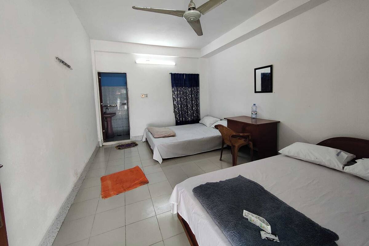 Ein Hotelzimmer mit zwei Betten, einem Schreibtisch, einem Stuhl, einem Deckenventilator, einem Badezimmer mit Waschbecken und einem Spiegel.