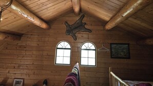 A log cabin room with a high ceiling, wooden beams, two windows, a hanging animal head, and a bed. - Camp Jackson Family Cabin (San Juan County)