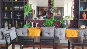 Ein modernes Wohnzimmer mit einer Couch, Sesseln und einem Esstisch. Ein großer Spiegel mit dekorativem Rahmen sowie Grünpflanzen sind zu sehen. Regale mit Gläsern und Büchern sind ebenfalls sichtbar.
