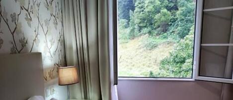 Una habitación con cama, mesita de noche, lámpara y ventana con vista a follaje.