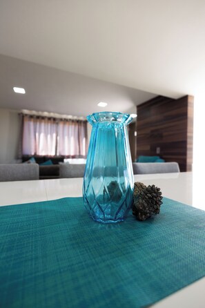 Un jarrón azul sobre una mesa blanca con un mantel individual teal con textura, una piña y un salón moderno de fondo con un sofá gris y un revestimiento de pared de madera.