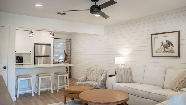 Een moderne keuken met witte kasten, een roestvrijstalen koelkast en hanglampen. Er is een eethoek met een witte tafel en twee krukken. Een grijze fauteuil en een beige hocker staan voor een witte bank.