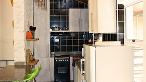 A compact kitchen with a stove, oven, and white refrigerator. There is a wooden shelf with kitchen utensils and a green chair.