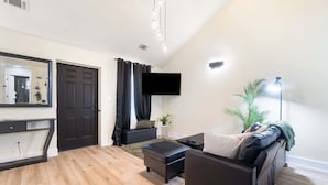A modern living room with a flat-screen TV, a sofa, a coffee table, and a dining area with a mirror and a door.