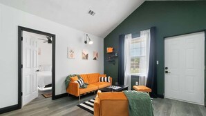 Ein modernes Wohnzimmer mit einer Couch, einem Couchtisch und einem kleinen Tisch mit einer Lampe. Zwei Türen, eine führt ins Schlafzimmer, die andere in den Flur.