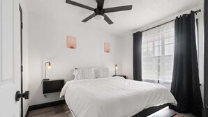 Ein Schlafzimmer mit einem Bett, Deckenventilator, Nachttischlampen, einem Fenster mit Vorhängen und Kunstwerken an der Wand.
