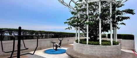 Zona de ejercicios al aire libre con elípticas y un espacio circular de descanso con un árbol.
