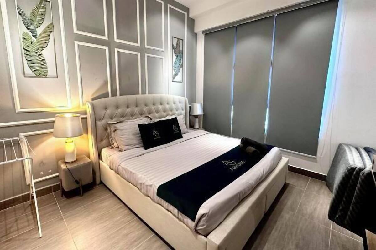 Un dormitorio moderno con cabecera capitonada, cama con sábanas blancas, una manta oscura y un ventanal con persianas.