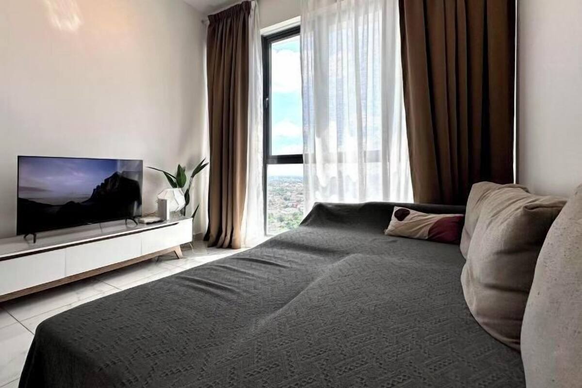 Una habitación de hotel moderna con una cama grande, un televisor de pantalla plana sobre un mueble blanco, vistas de la ciudad a través de un ventanal amplio y una planta en maceta.