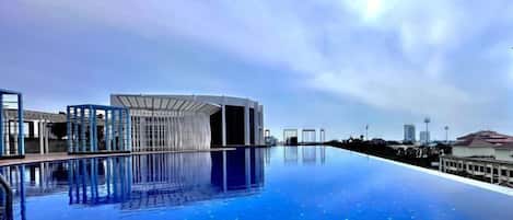 A modern hotel with a large infinity pool reflecting the sky, a contemporary building structure, and a clear view of the city skyline in the background.
