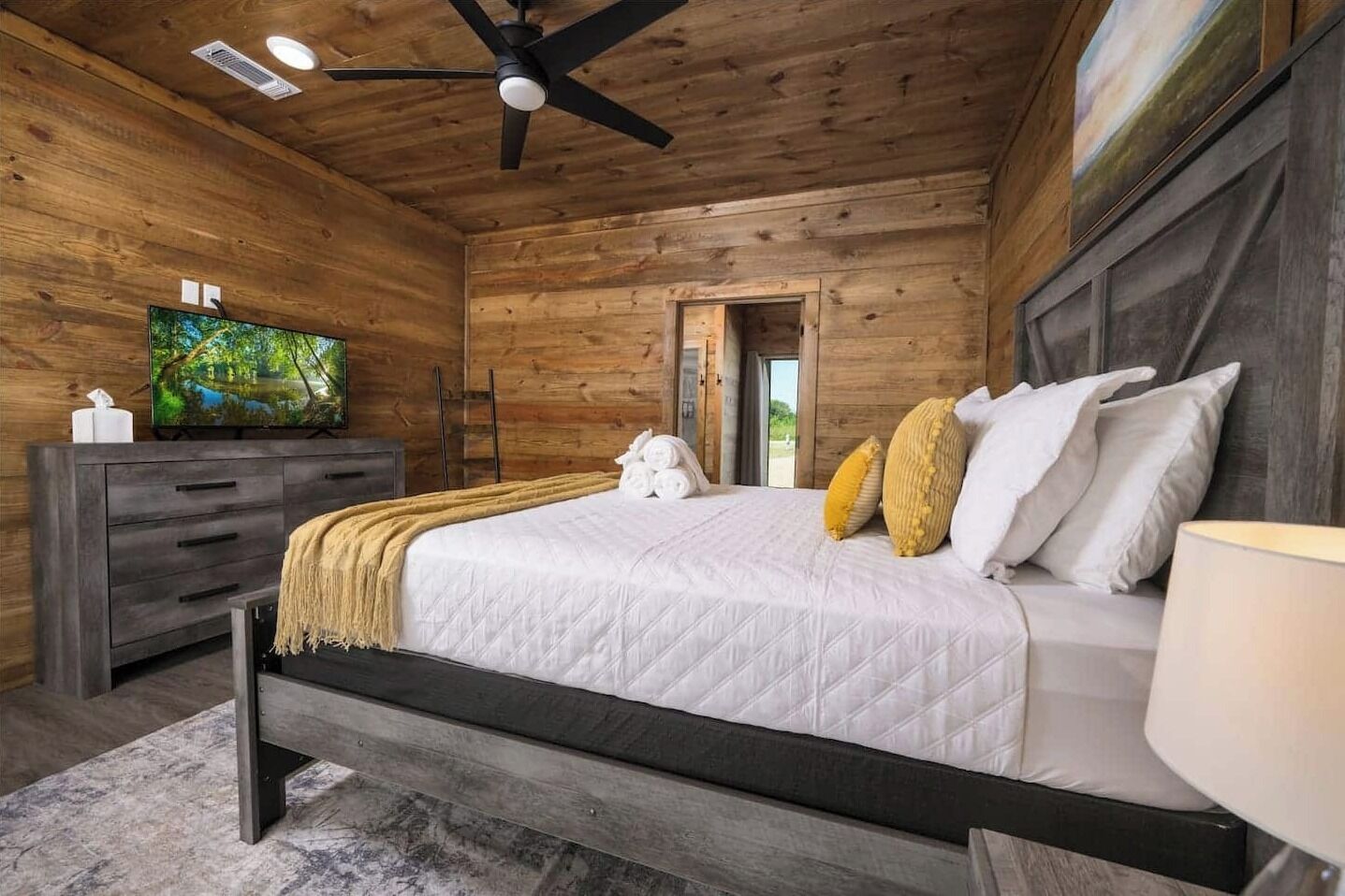 Quarto com teto de madeira, cama com roupa de cama branca, televisão sobre uma cômoda e porta que dá para o exterior.