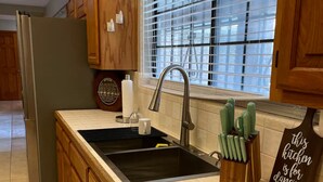 Una cocina con gabinetes de madera, un lavamanos de acero inoxidable, lavavajillas empotrado y una ventana con persianas.