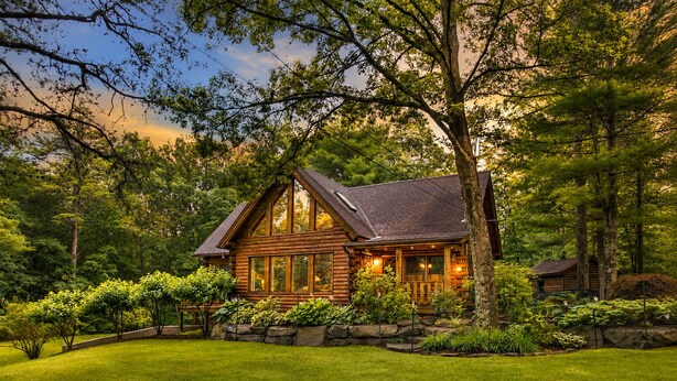 Une cabane en rondins dotée d’une grande véranda, entourée d’une végétation luxuriante et d’arbres.