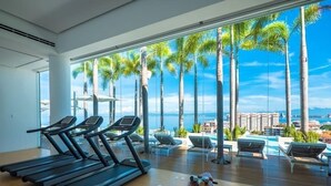 Ein Fitnessraum mit Laufbändern und einer Bank, mit Blick auf einen Pool mit Palmen und Meerblick.