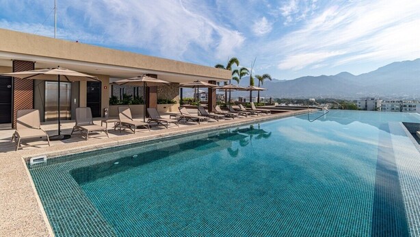 Ein Rooftop-Infinity-Pool mit Liegestühlen und Sonnenschirmen mit Blick auf eine Stadtlandschaft und Berge.