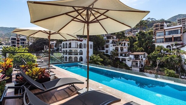 A rooftop pool area with lounge chairs and umbrellas, surrounded by white buildings with balconies and greenery.