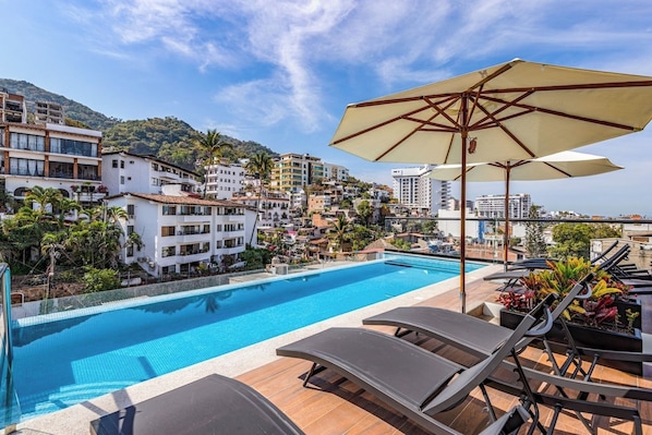 A rooftop pool with lounge chairs and umbrellas, surrounded by buildings and a hillside.