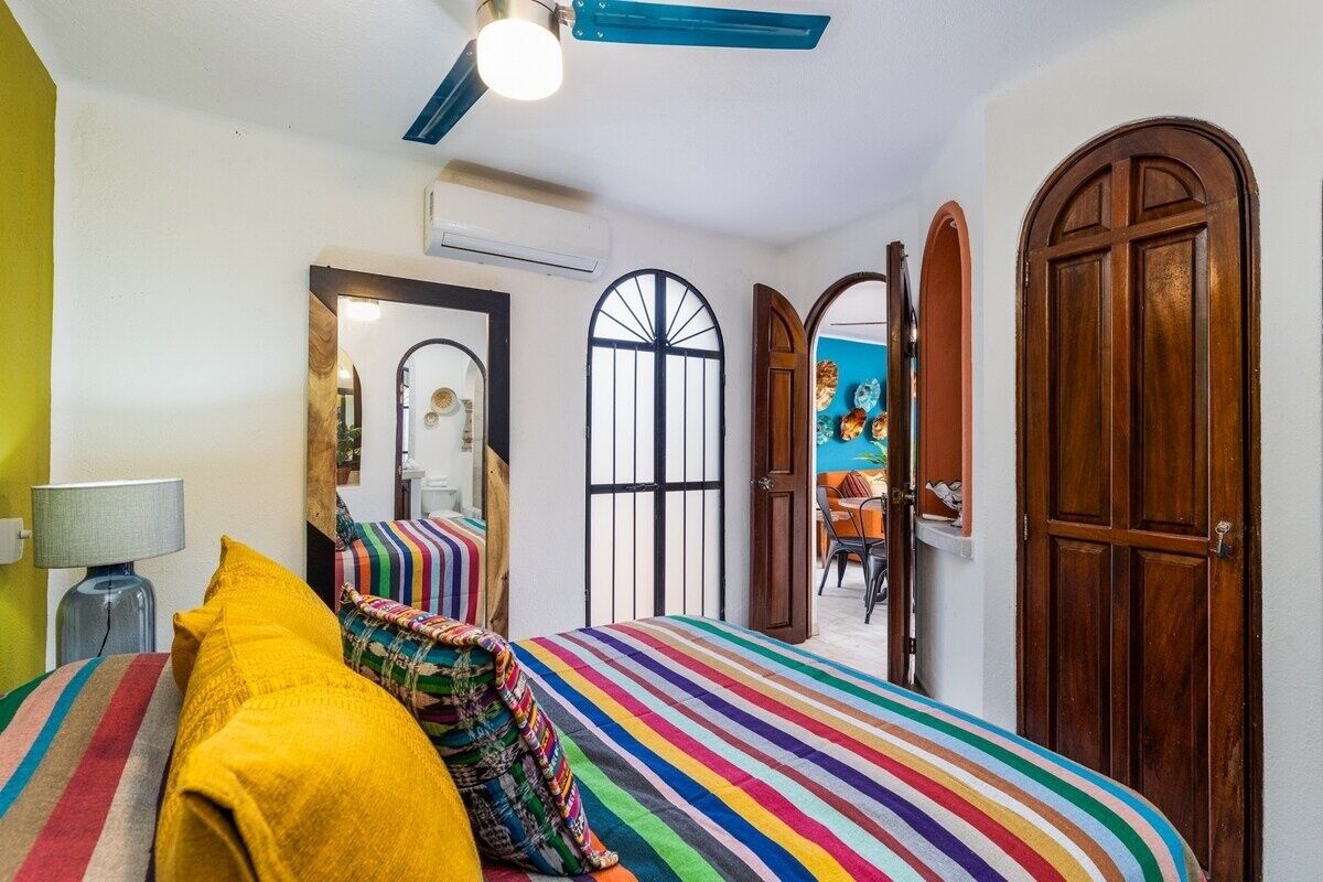 Ein Schlafzimmer mit gestreifter Bettdecke, Deckenventilator, Spiegel und Blick auf den Essbereich durch eine geöffnete Tür.