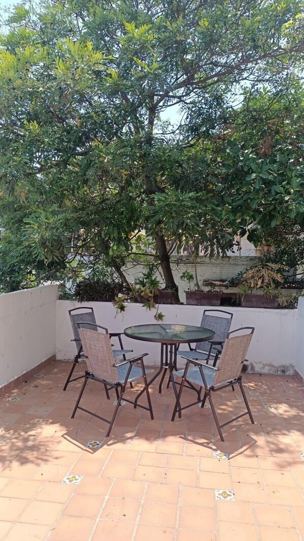 Un patio al aire libre con una mesa redonda y seis sillas, rodeado por un muro blanco y un árbol grande que proporciona sombra.