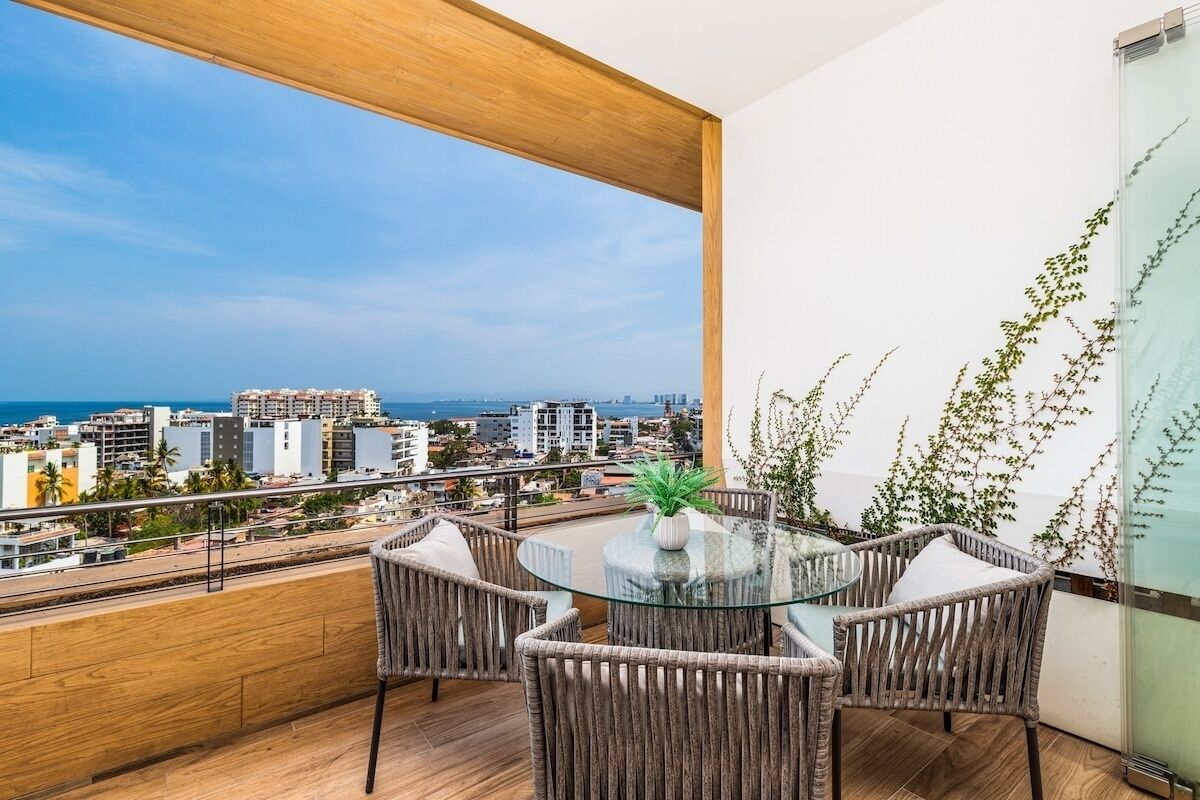 Ein Balkon mit Korbmöbeln, einem Glastisch und Blick auf Stadt und Meer.