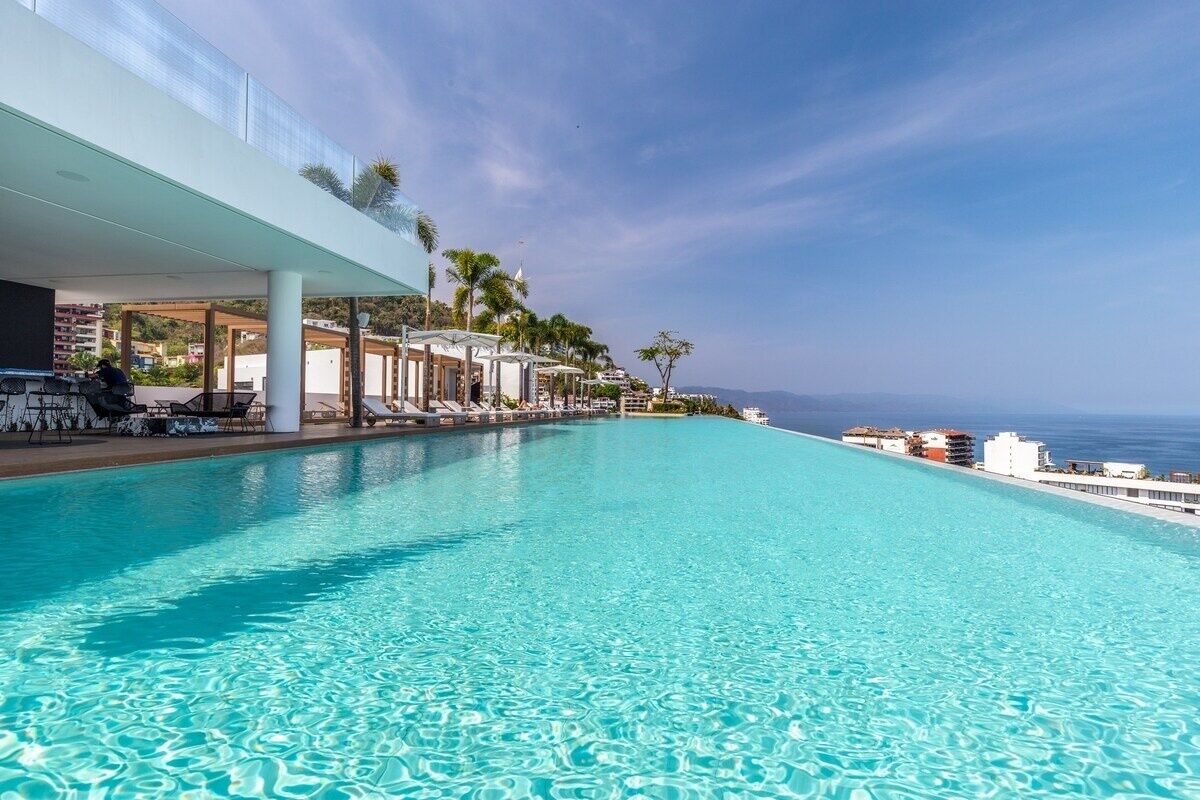 A clear blue infinity pool with lounge chairs and umbrellas, overlooking a coastal area.