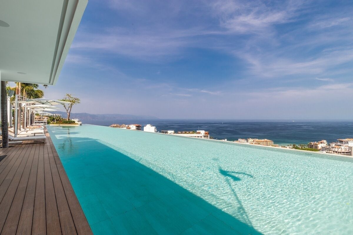 Piscina infinita de color azul cristalino con terraza de madera, con vistas a una playa y al océano.