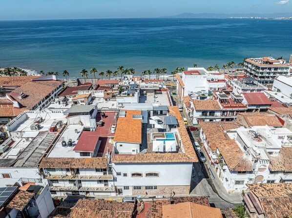 An aerial view of a coastal urban area with buildings, some with red-tiled roofs, and a clear view of the ocean.