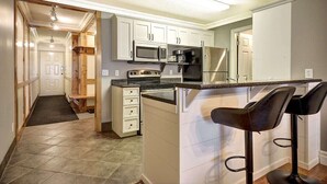 A kitchen with white cabinets, a microwave, a stove, a refrigerator, and a bar area with stools. - The Eagles at Big White (Beaverdell)