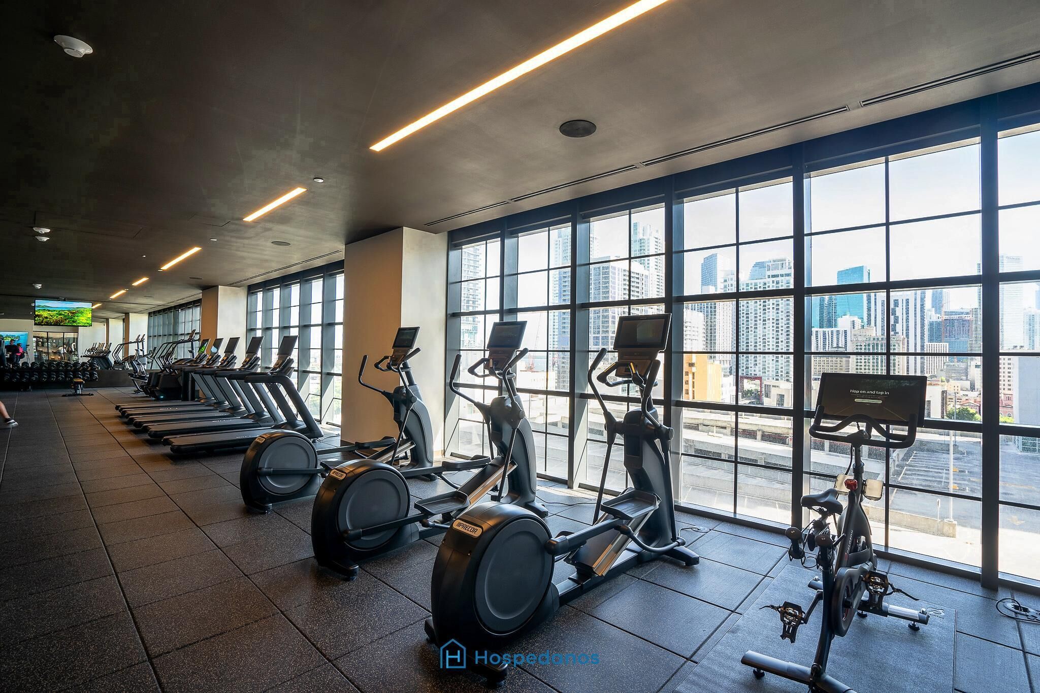 A gym with treadmills, elliptical machines, and a city view through large windows.