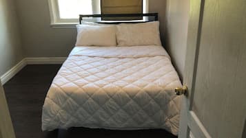 Una cama pequeña y ordenada con una colcha blanca acolchada y dos almohadas, ubicada en una habitación con ventana y puerta.