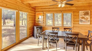Interno di una baita in legno con un tavolo da pranzo, sedie, una ventola a soffitto e ampie finestre che si affacciano sulla foresta.