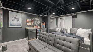 A modern, well-lit room with a grey sofa, a bench, a small kitchenette, and a door leading to another room. - Live the Game: Pool, Cinema, Gym, Jersey Wall (Massillon)